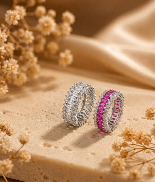 Two silver rings with rectangular gemstones, one silver and one pink, displayed on a beige stone surface with dried flowers and draped satin fabric.