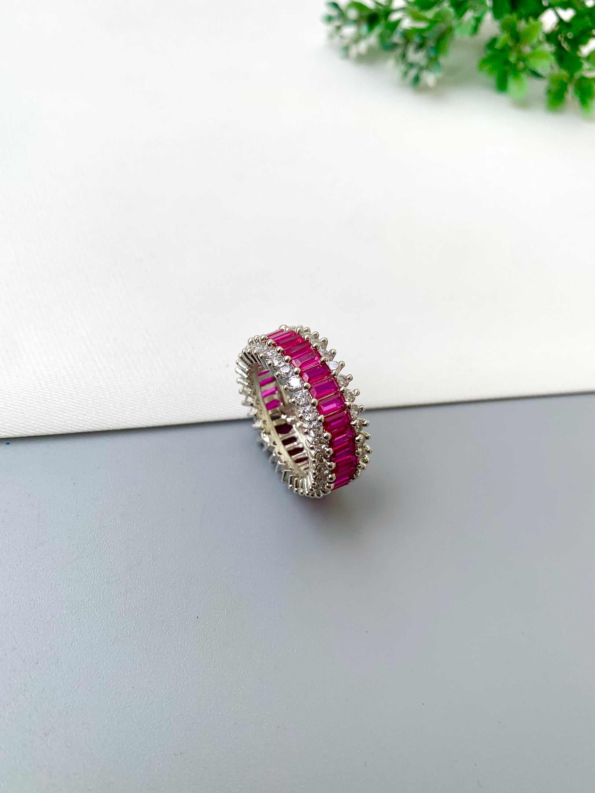 A silver-toned eternity ring featuring two rows of vibrant pink gemstones and clear accent stones, displayed on a split white and gray surface with a small green plant in the background.