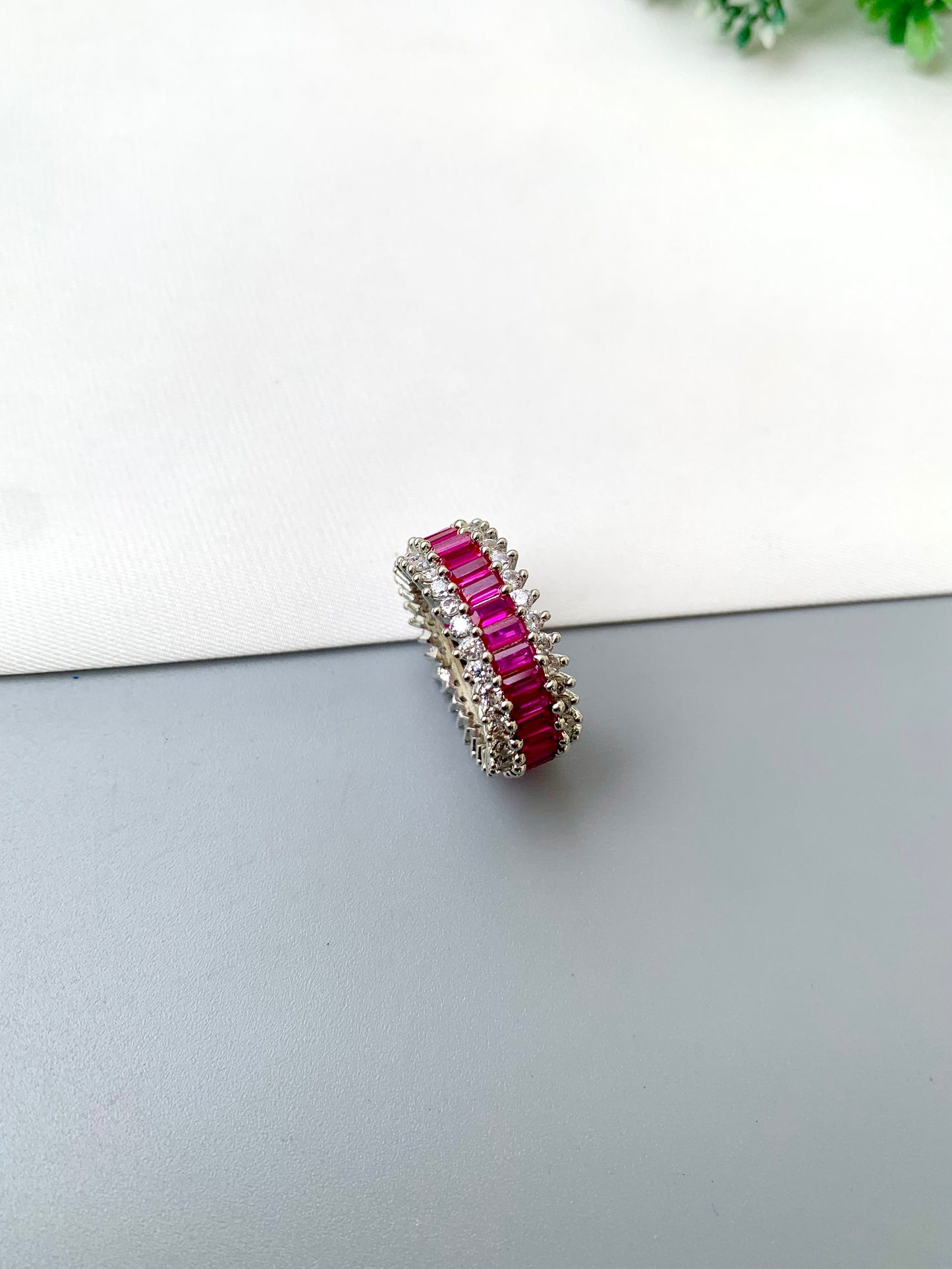 A silver-toned ring featuring a vertical channel setting of bright pink baguette-cut gemstones, photographed on a split white and gray surface.
