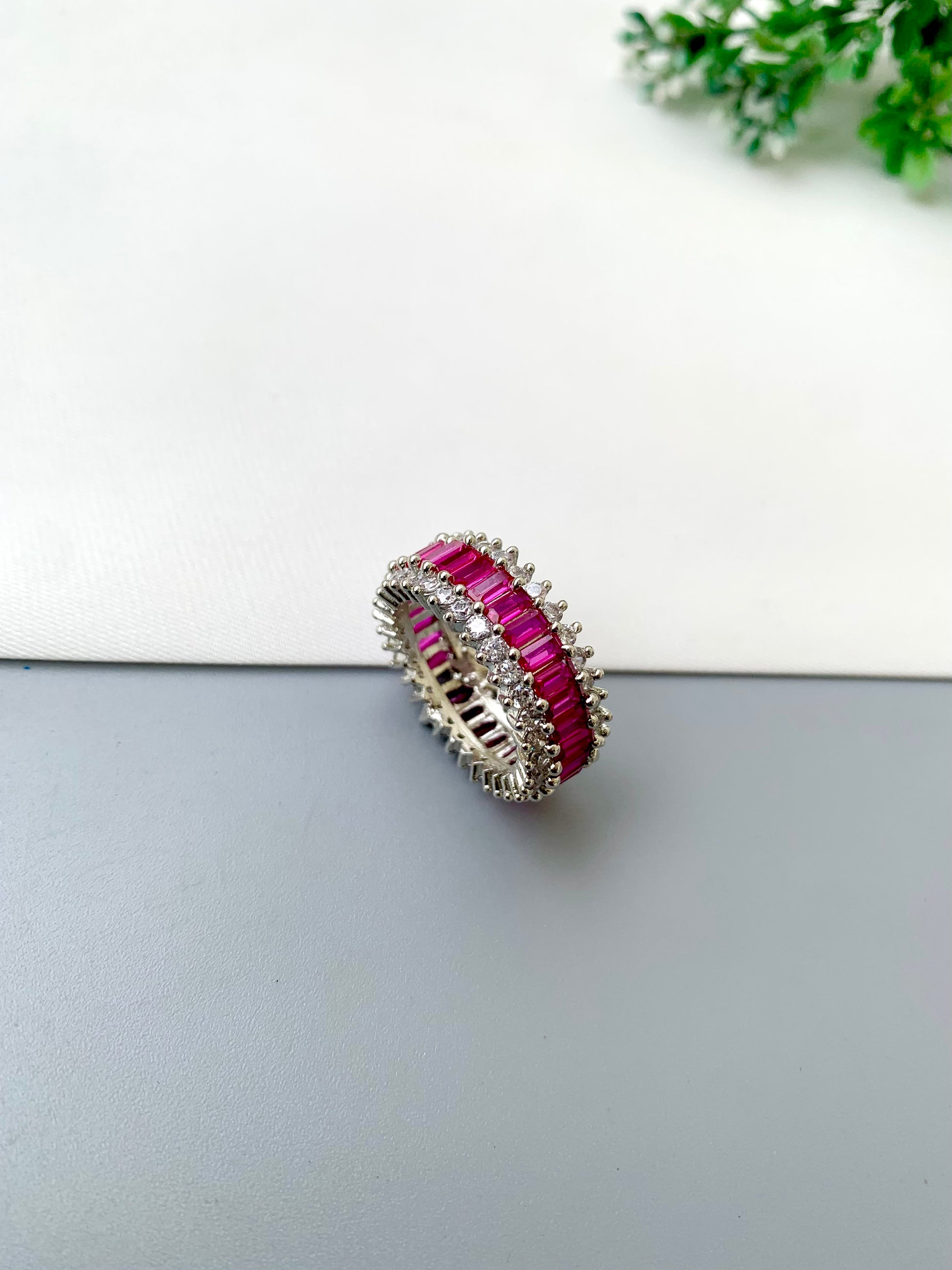 A silver-toned eternity ring featuring rows of vibrant pink baguette-cut gemstones and clear accent stones, displayed on a split white and gray surface with a small green plant in the corner.