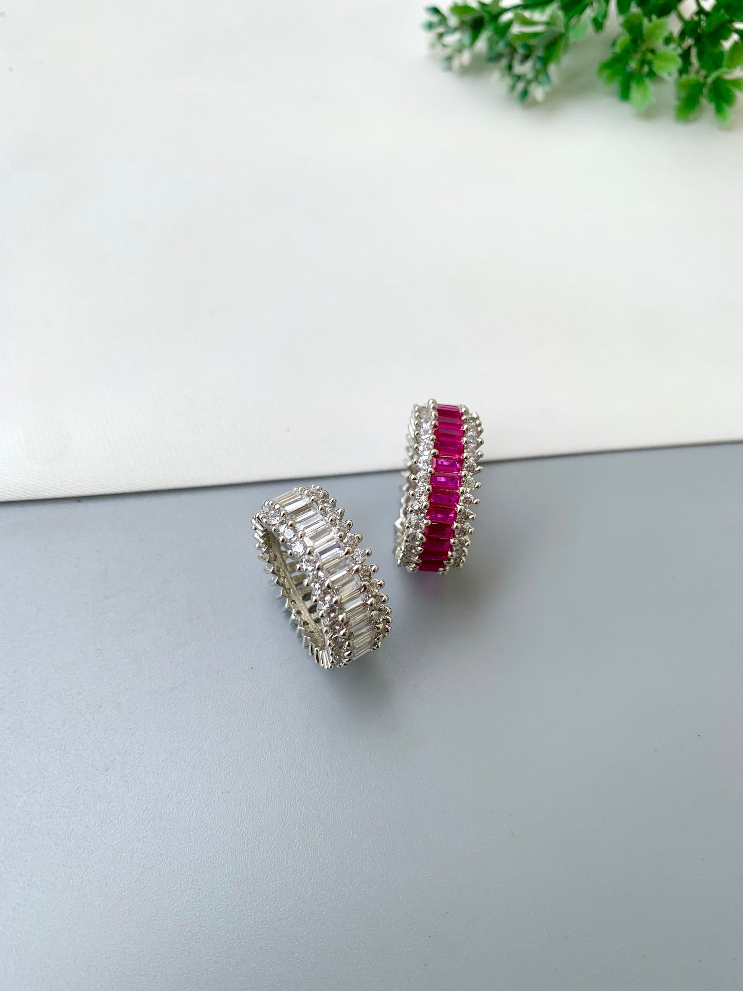 A pair of silver-toned band rings with a central row of bright pink rectangular gemstones, photographed on a split white and gray surface with a small green plant in the corner.