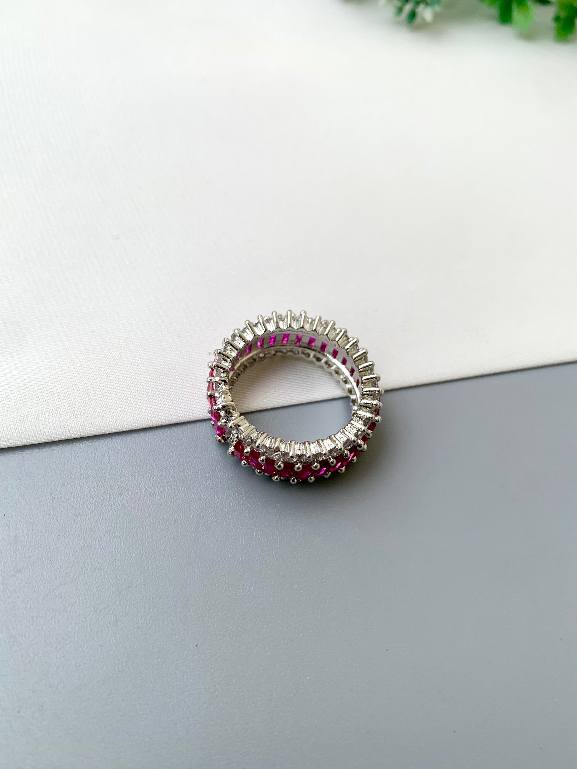 A silver-toned ring featuring rows of small clear stones and alternating rows of deep pinkish-red gemstones, photographed on a split white and gray surface.