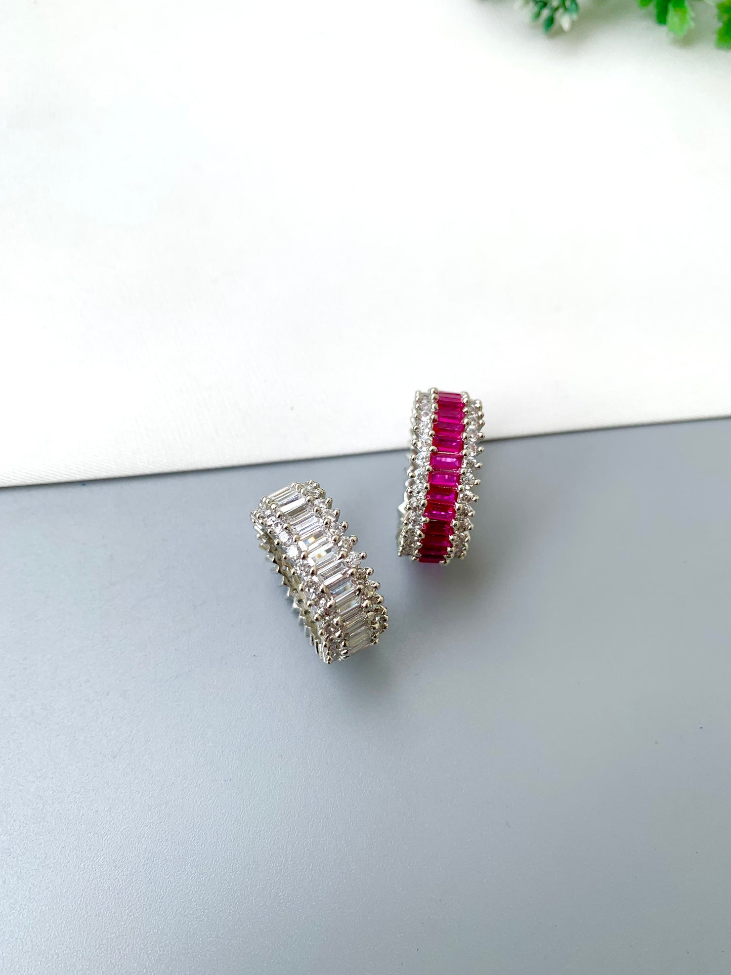 A pair of silver-toned cylindrical earrings with clear baguette-cut stones and one earring featuring a row of bright pink gemstones, displayed on a split white and gray surface.