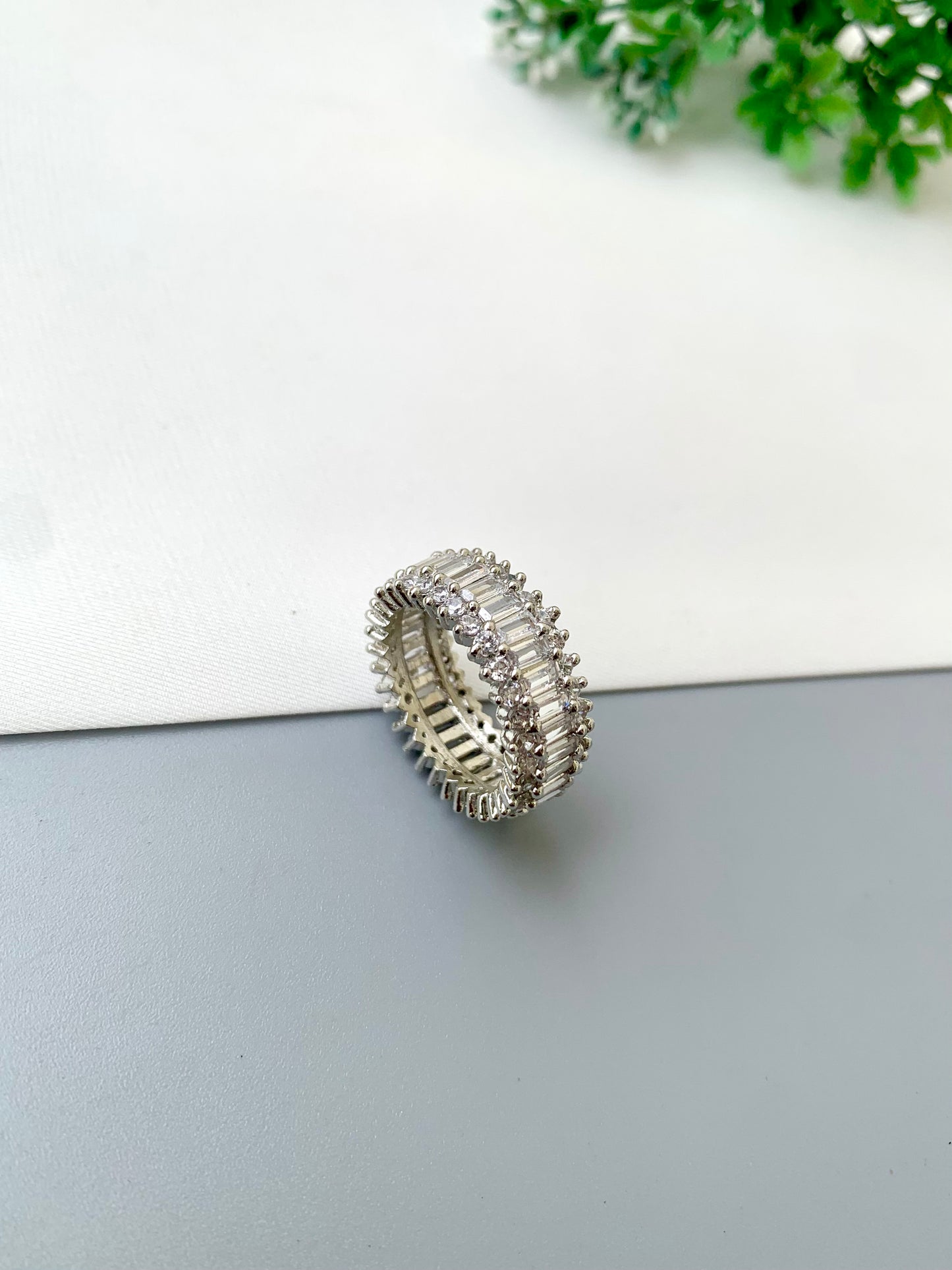 A silver-toned eternity ring set with clear baguette and round stones, resting on a split white and gray surface with a small green plant in the corner.