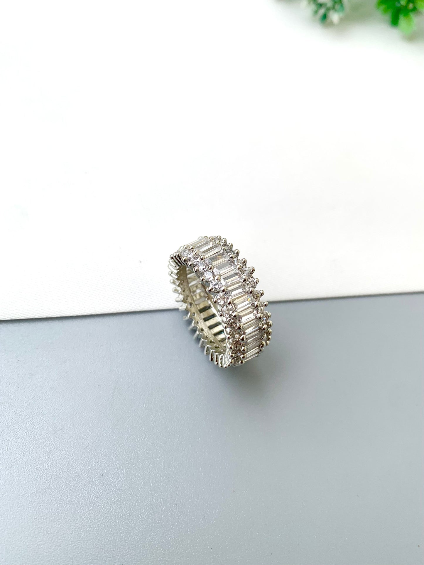 A silver band ring embellished with multiple rows of clear baguette and round-cut stones, photographed on a split white and gray surface.
