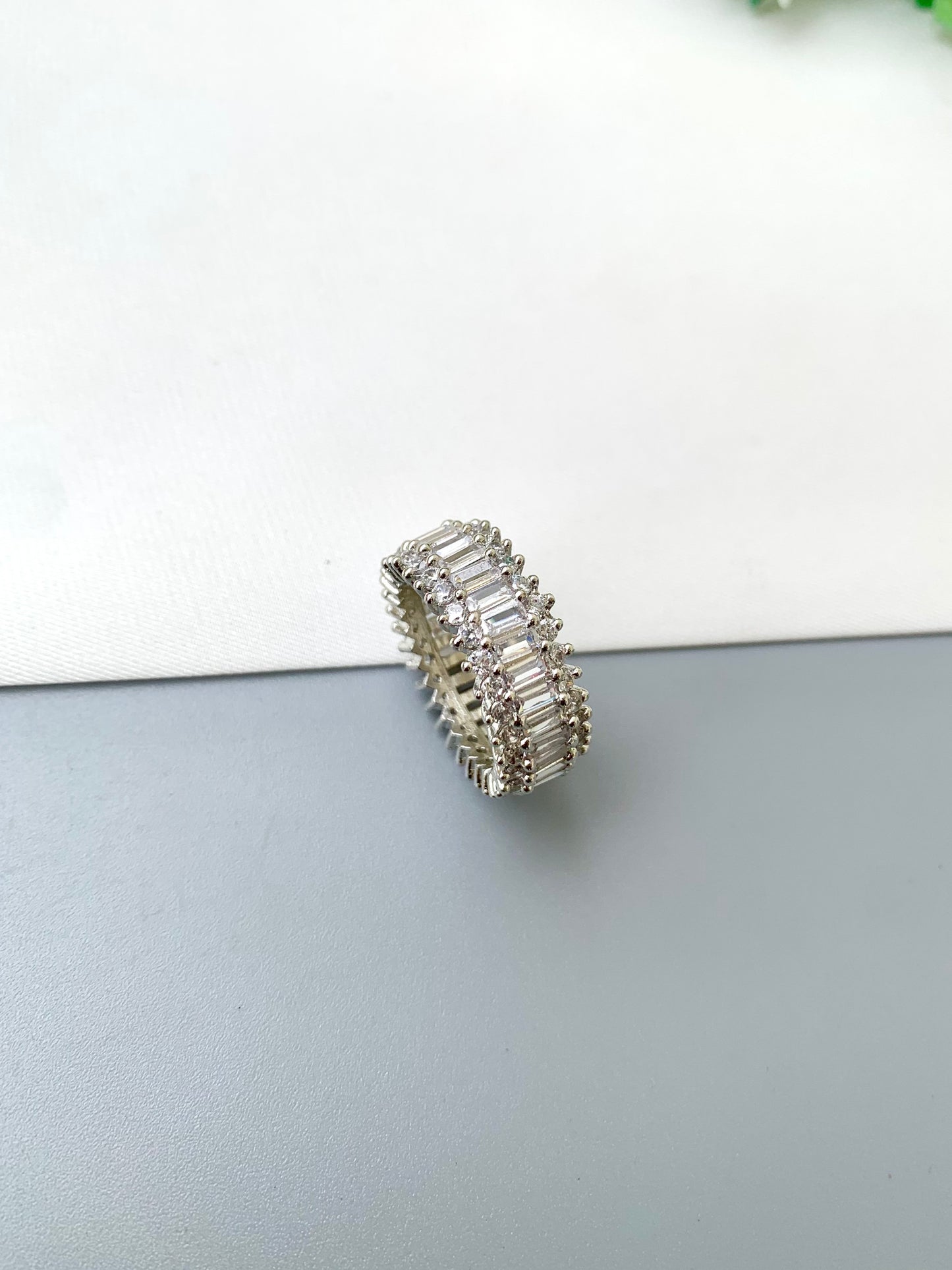 A silver band ring set with multiple rows of baguette-cut clear stones, photographed on a split white and gray surface.