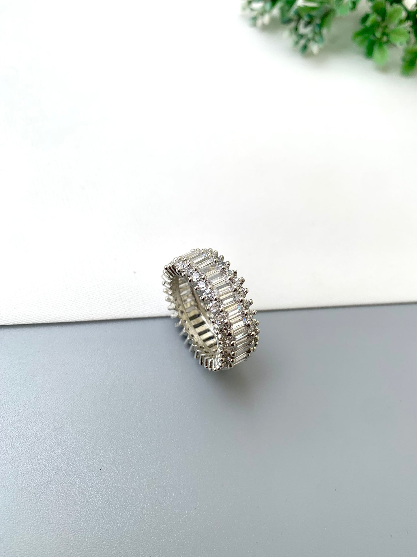A silver-toned eternity ring set with baguette-cut clear stones, resting on a split white and gray surface with a small green plant in the background.