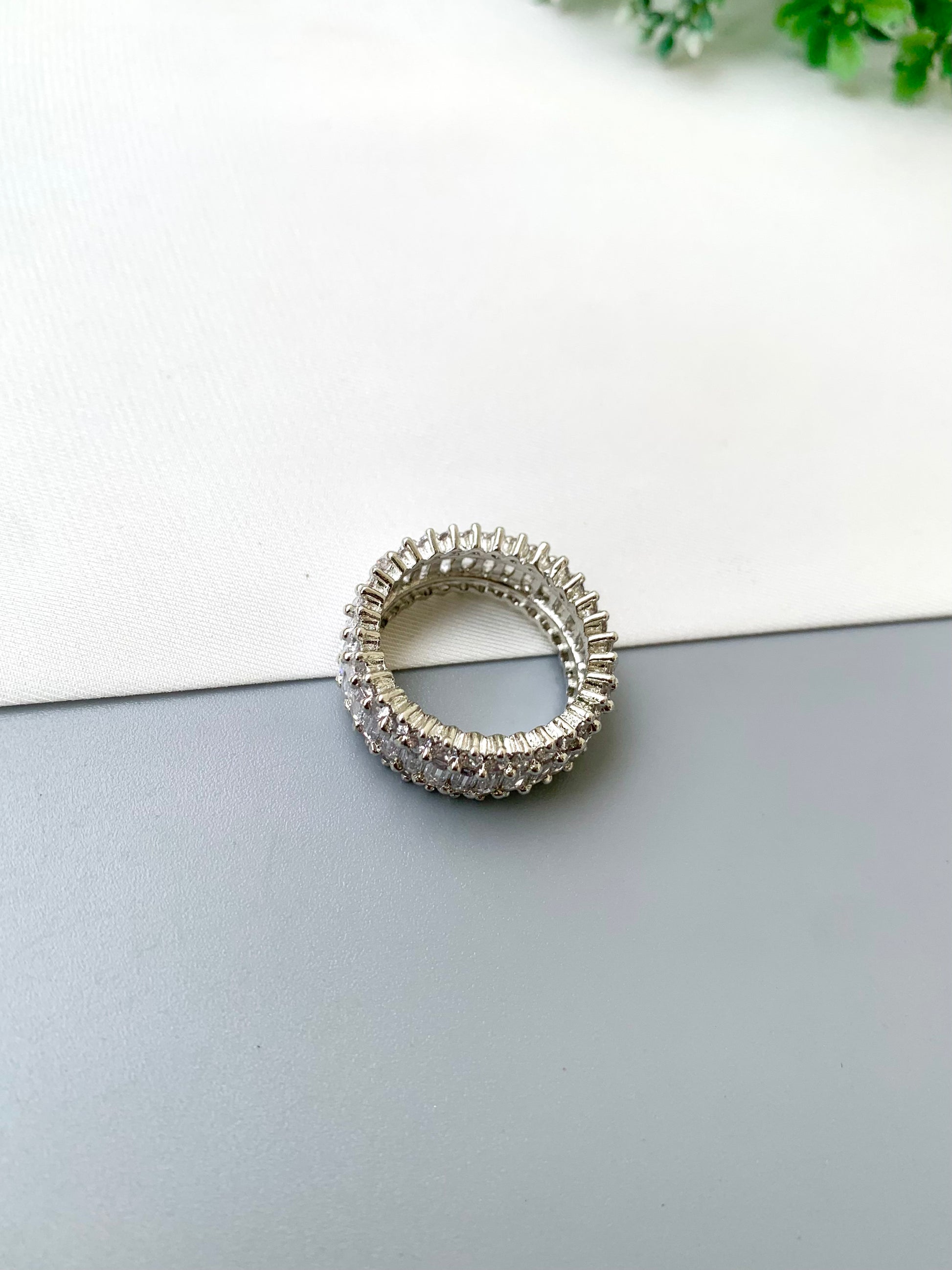 A silver-toned tennis-style ring with clear stones, photographed on a split white and gray surface with a small green plant in the corner.