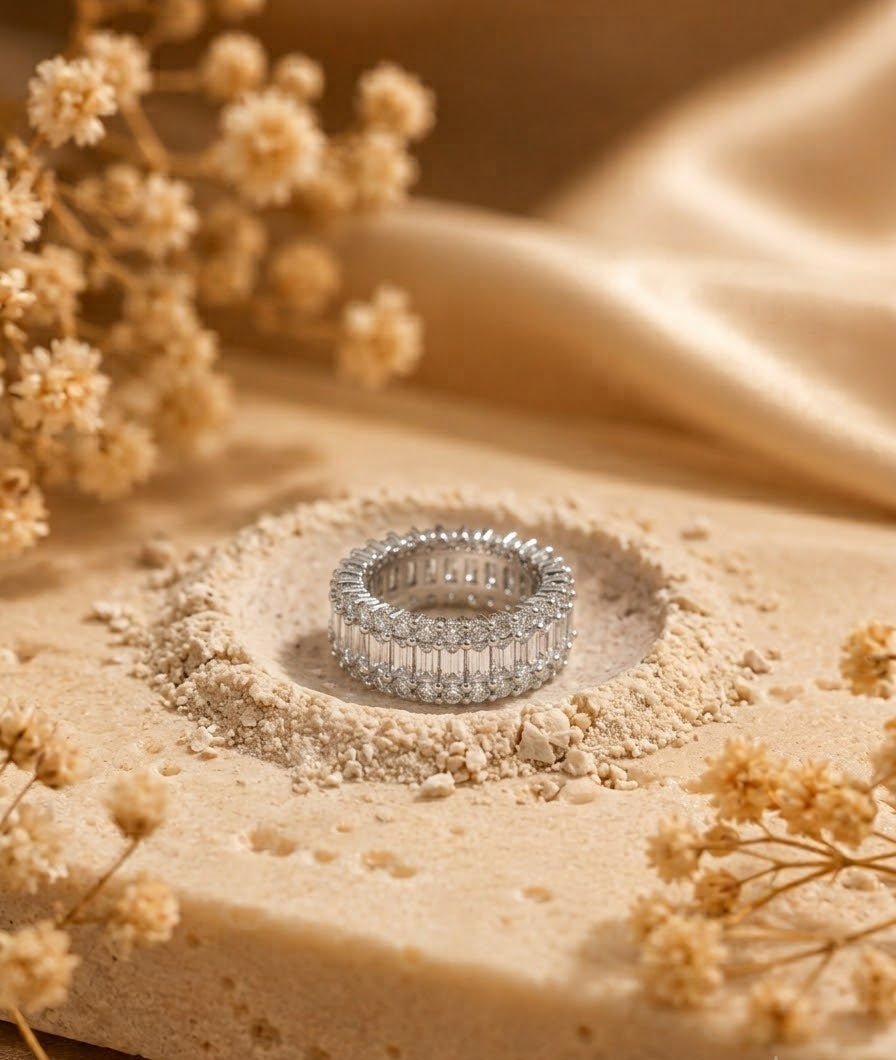 A silver eternity ring set with clear baguette and round stones, displayed in a shallow pit of beige powder with dried flowers and draped satin fabric in the background.