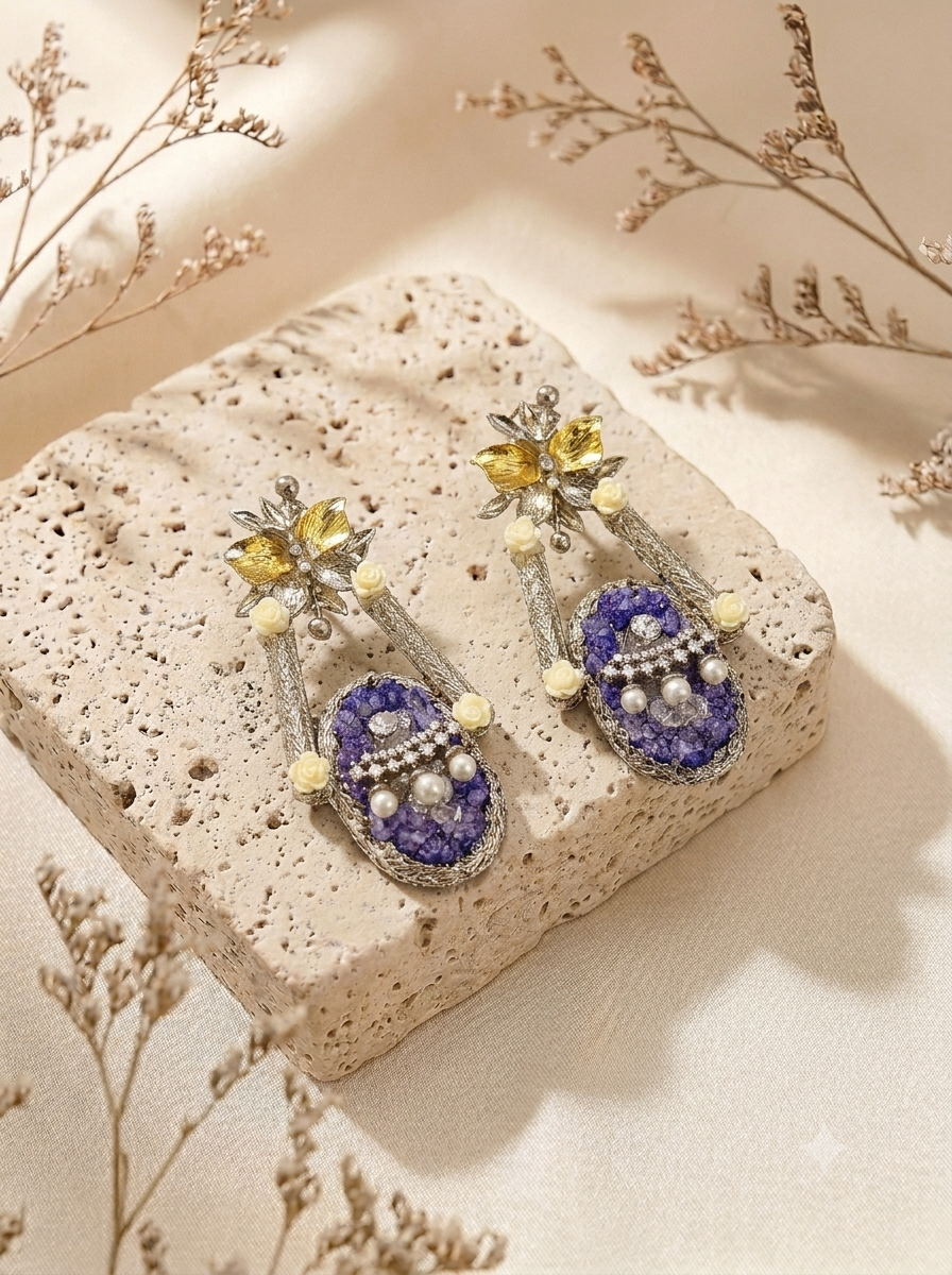 A pair of ornate drop earrings displayed on a textured beige stone slab. The earrings feature oval purple and blue stone drops with silver-toned settings, small cream pearl-like accents, and floral tops with yellow gemstones.