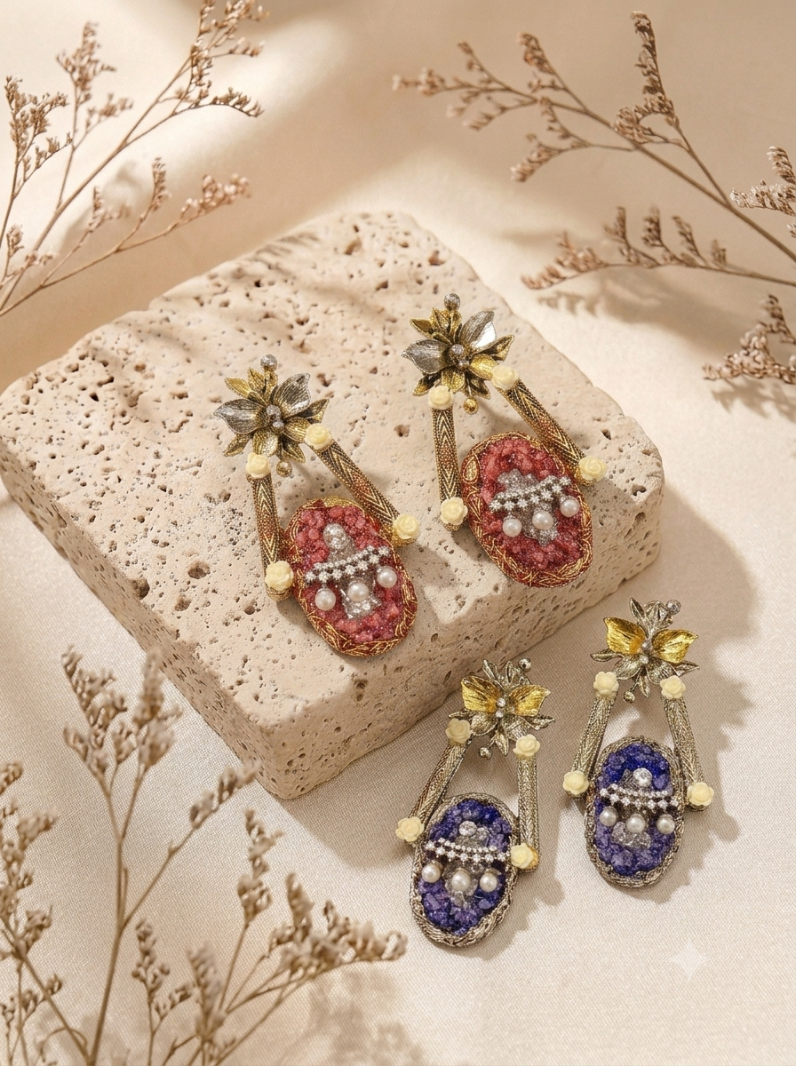 Two pairs of ornate drop earrings displayed on a textured stone slab and neutral surface. One pair features pink enamel with silver and pearl-like details, and the other pair features deep blue enamel with similar metallic and pearl-like embellishments.
