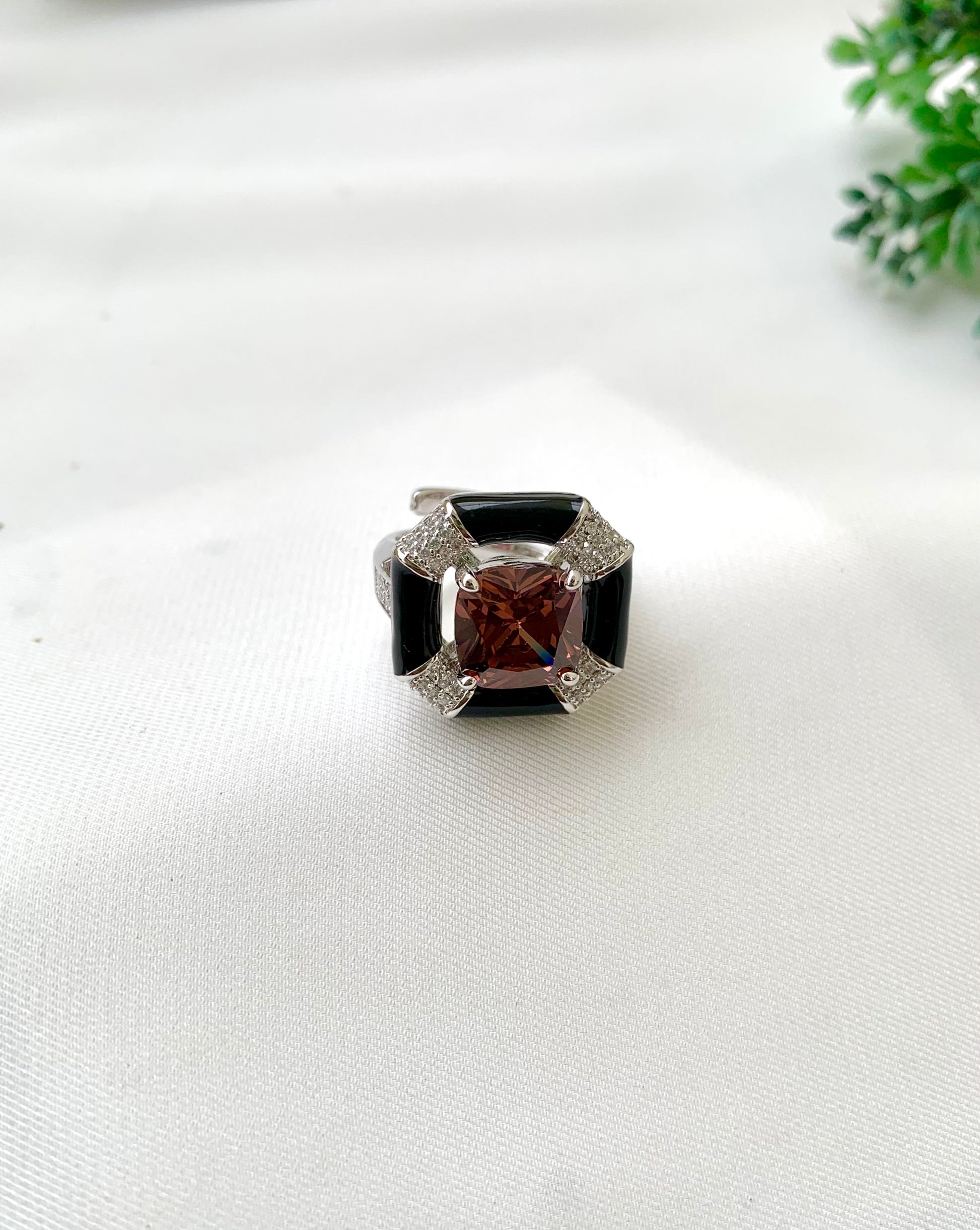 A square ring featuring a central cushion-cut reddish-brown gemstone surrounded by a black inlay and silver-toned metal with small clear stones, displayed on a white textured fabric background.