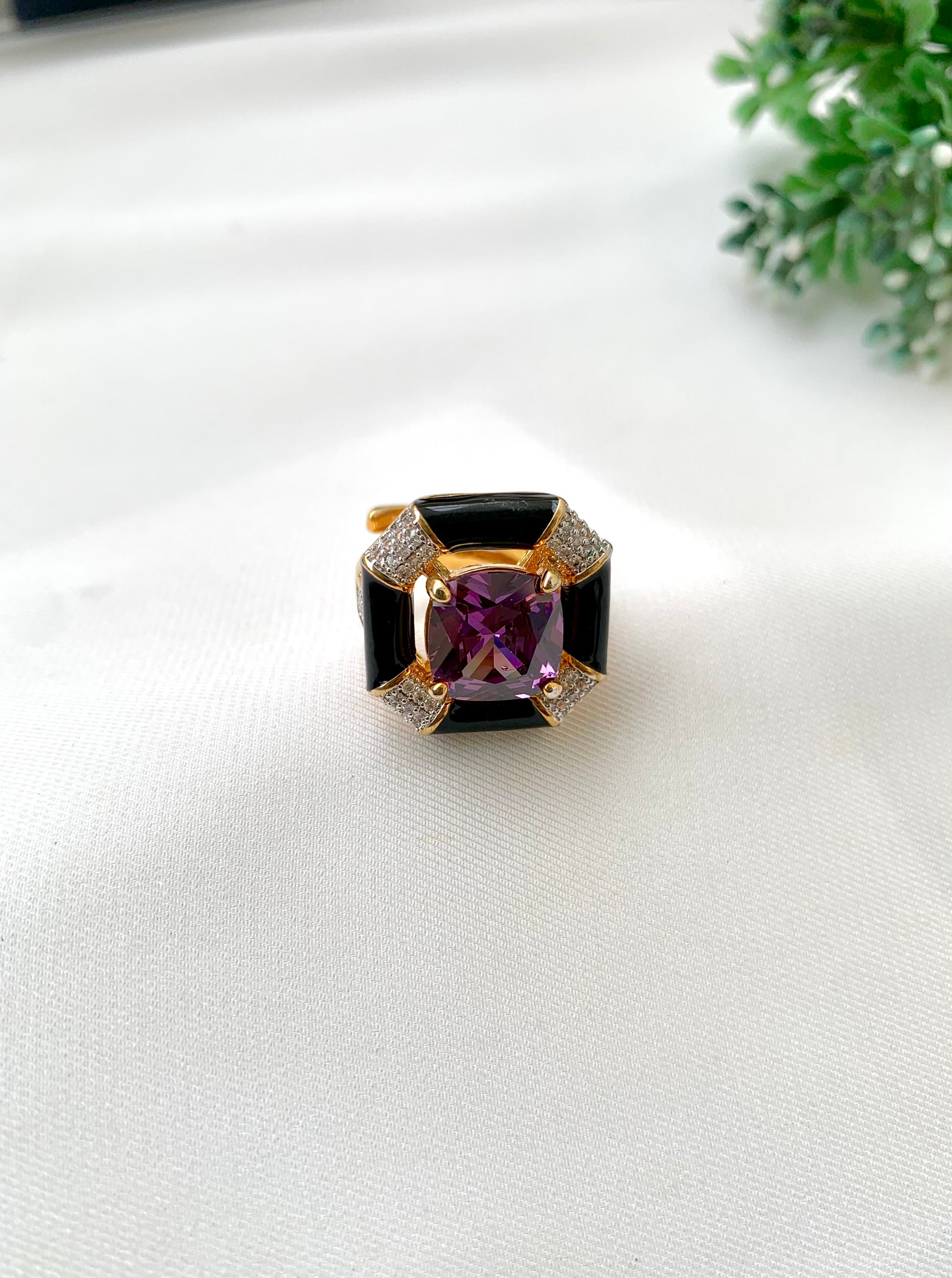 A square ring featuring a central cushion-cut purple gemstone, surrounded by black inlay and gold-toned metal with small clear stones, resting on a white textured fabric surface.