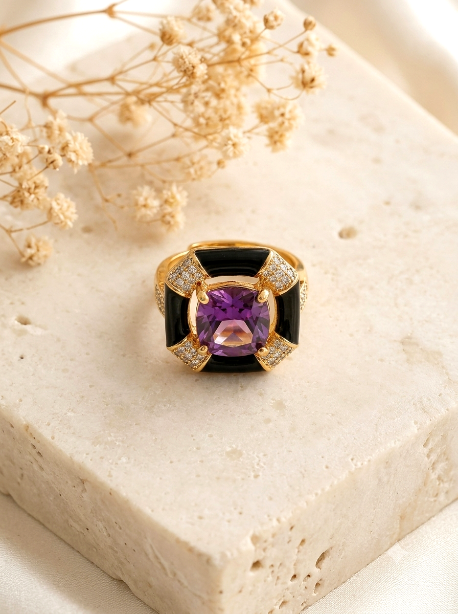 A gold ring with a large purple cushion-cut center stone and black inlay panels, accented with small clear stones, displayed on a beige stone slab with dried flowers.