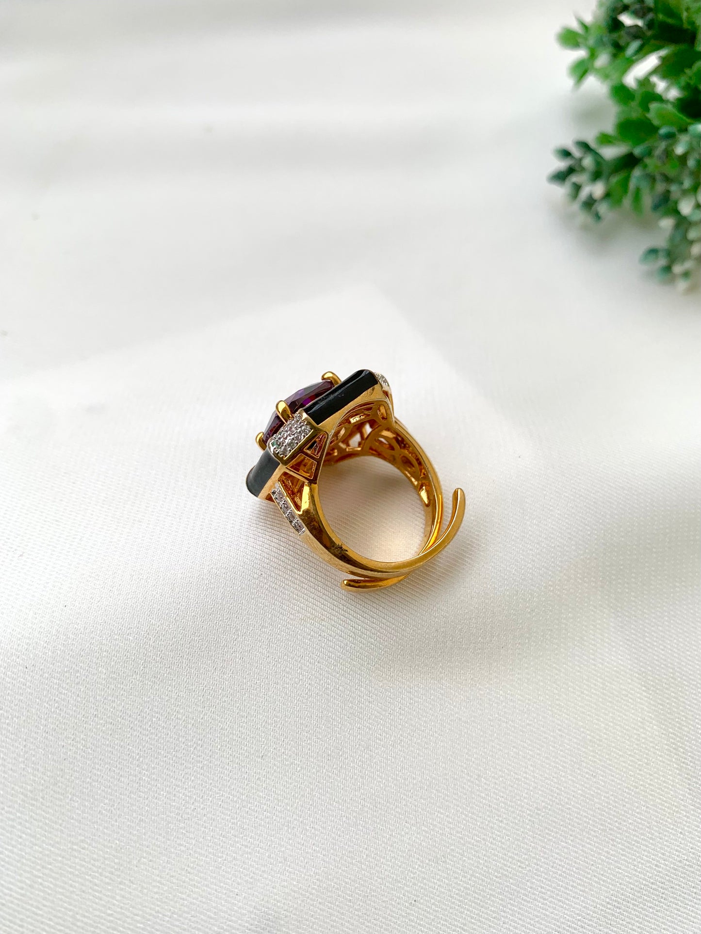 A gold ring with a dark red oval gemstone and small clear accent stones, displayed on a white textured fabric surface with a small green plant in the background.