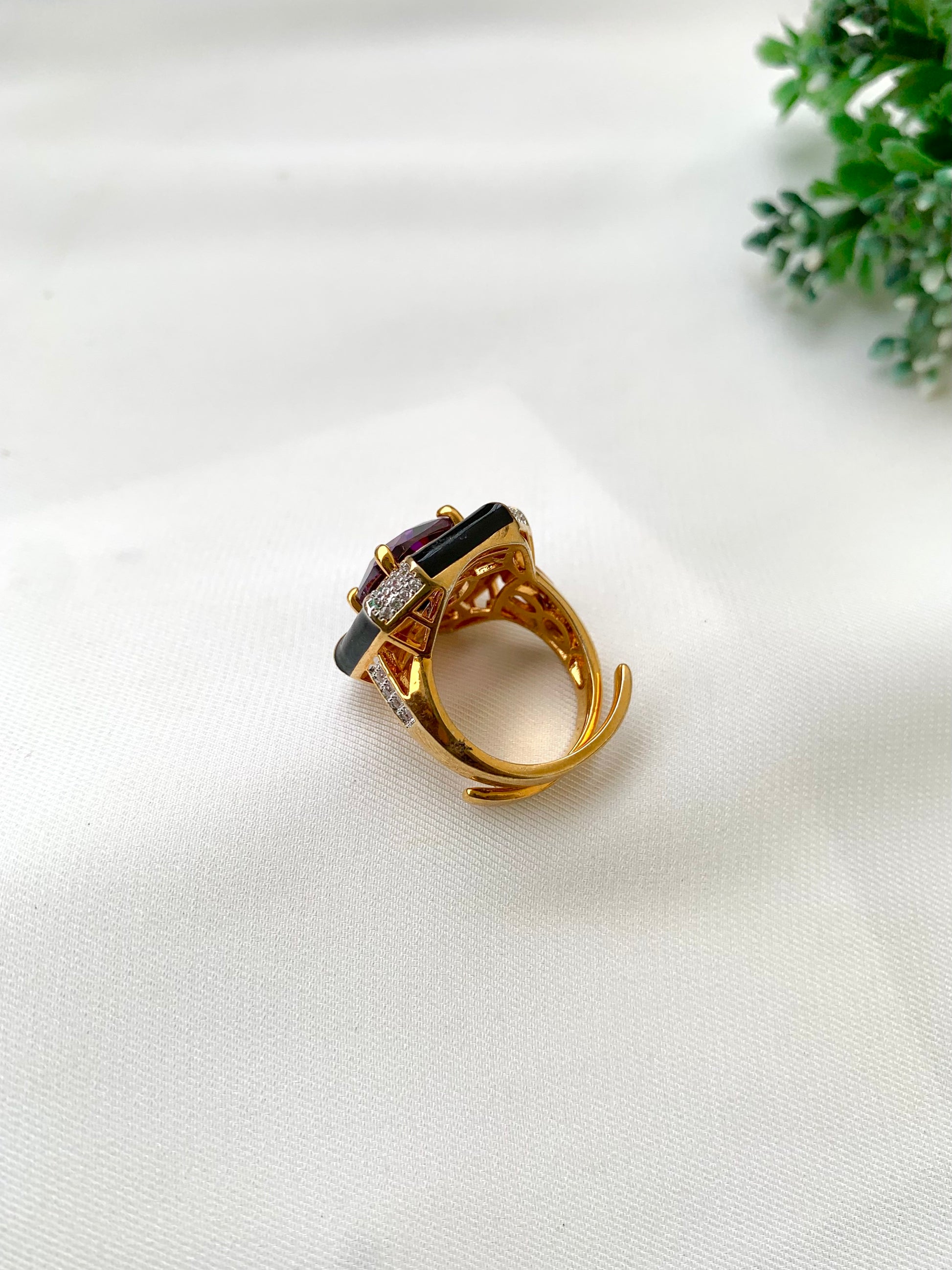 A gold ring with a dark red oval gemstone and small clear accent stones, displayed on a white textured fabric surface with a small green plant in the background.