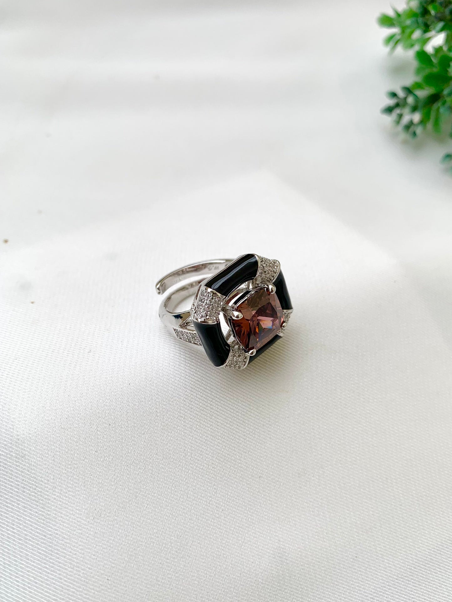 A silver-toned ring with a central cushion-cut brown gemstone, flanked by black inlay panels and pavé-set clear stones, resting on a white textured fabric surface.