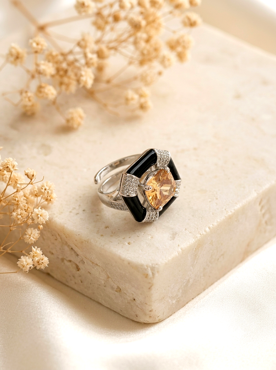 A silver ring with a large square-cut peach gemstone, black inlay, and pavé-set clear stones, displayed on a beige stone block with dried flowers.