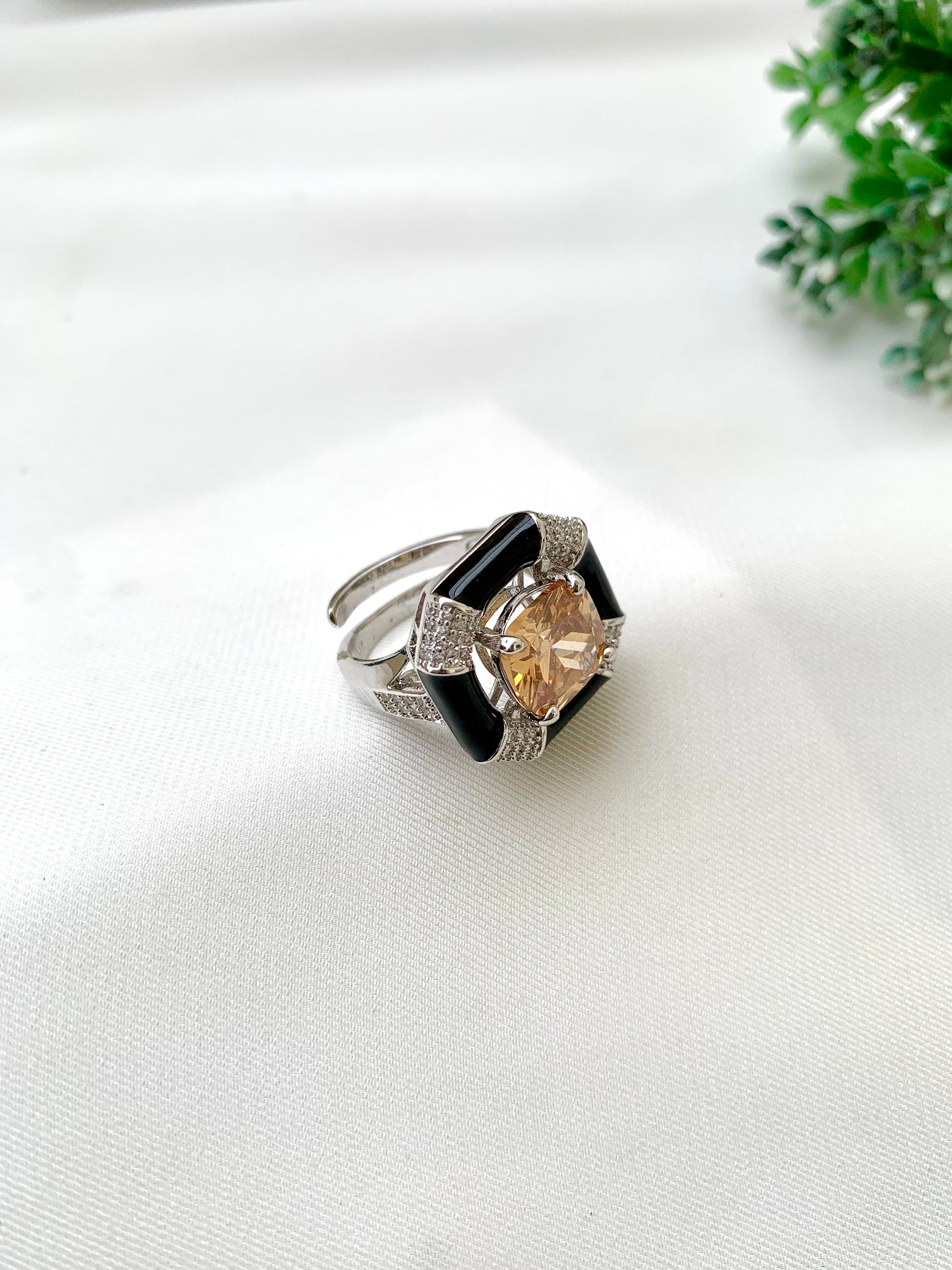 A silver-toned Art Deco style ring featuring a central peach-colored cushion-cut gemstone, surrounded by a black enamel inlay and pavé-set clear stones, resting on a white textured fabric surface.
