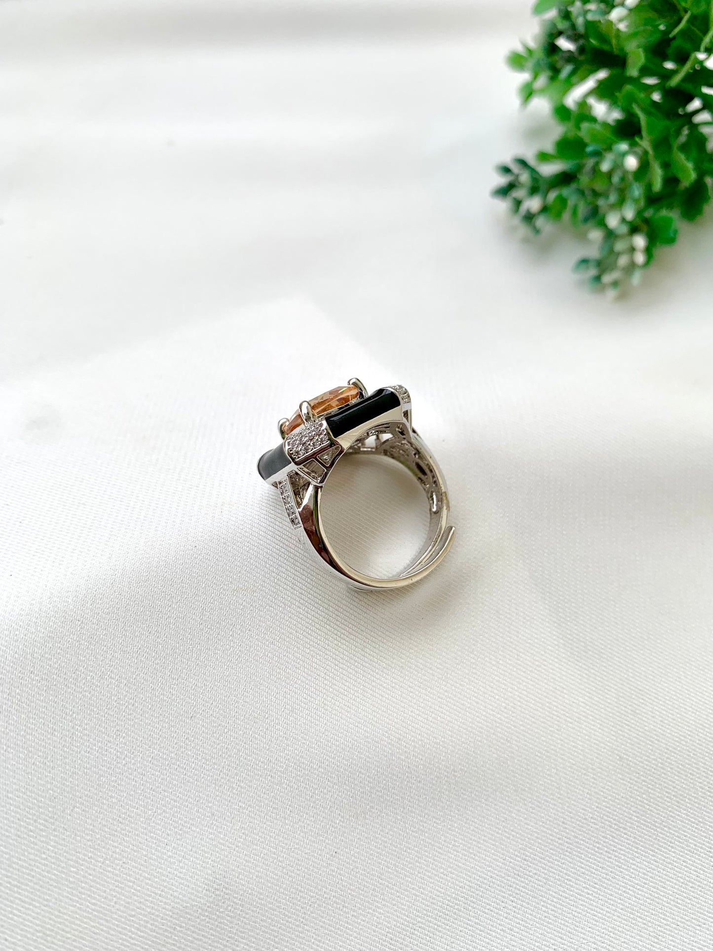 A silver-toned ring featuring a rectangular orange stone, a central black stone, and small clear accent stones, displayed on a white textured fabric background with a green plant in the corner.