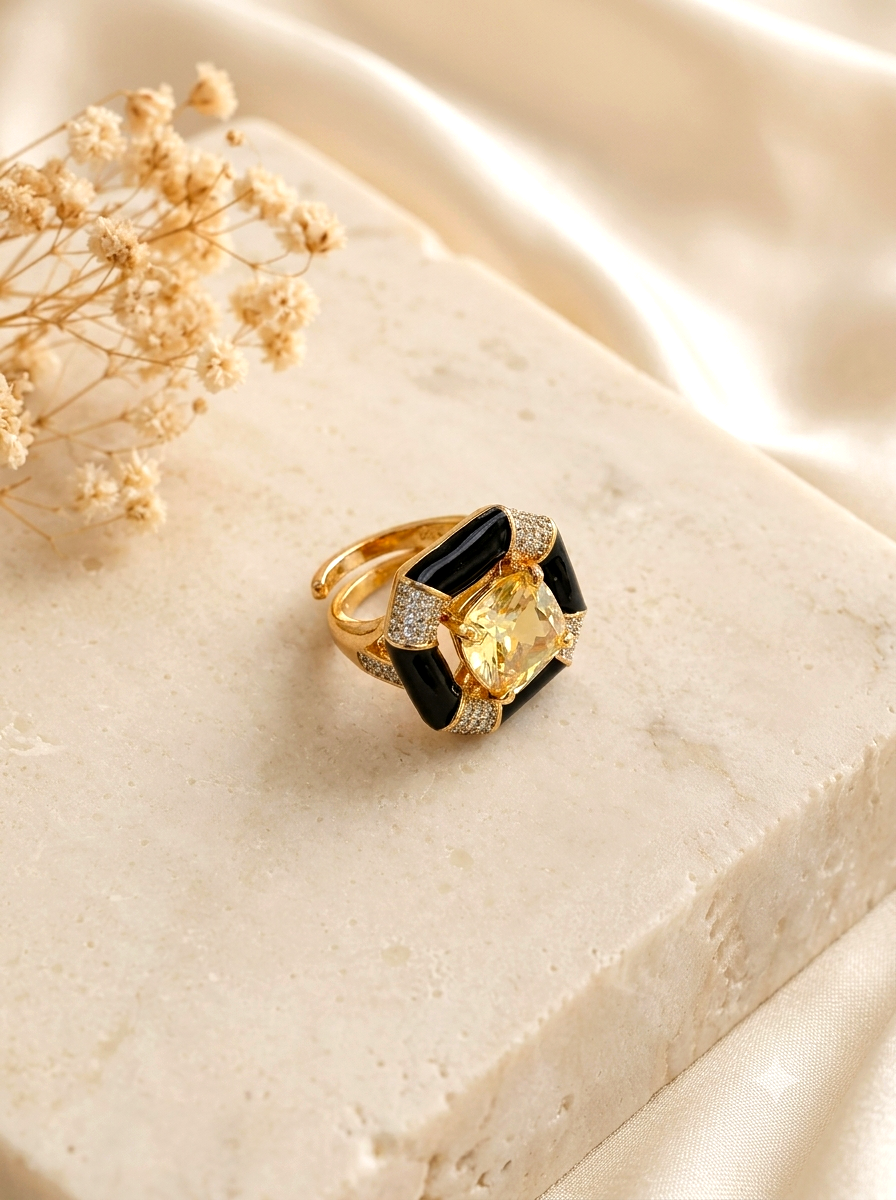 A gold ring with a large yellow cushion-cut center stone, black rectangular side stones, and small clear pavé accents, displayed on a cream stone slab with dried flowers and satin fabric.