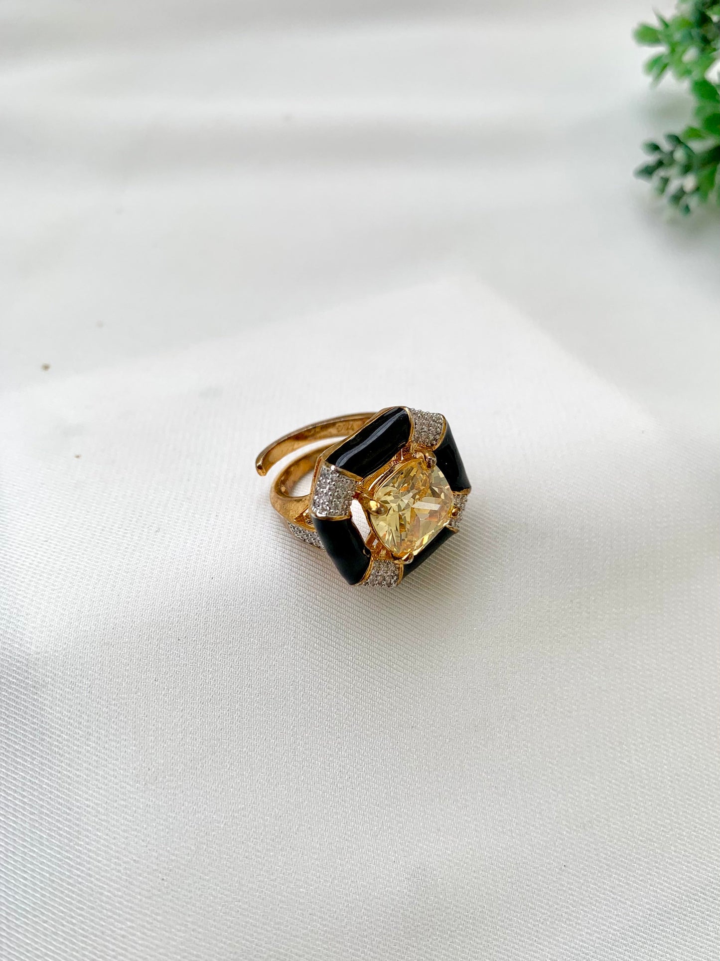 A gold ring with a large yellow cushion-cut center stone and black onyx geometric side stones, accented with small clear pavé stones, resting on a white textured fabric surface.
