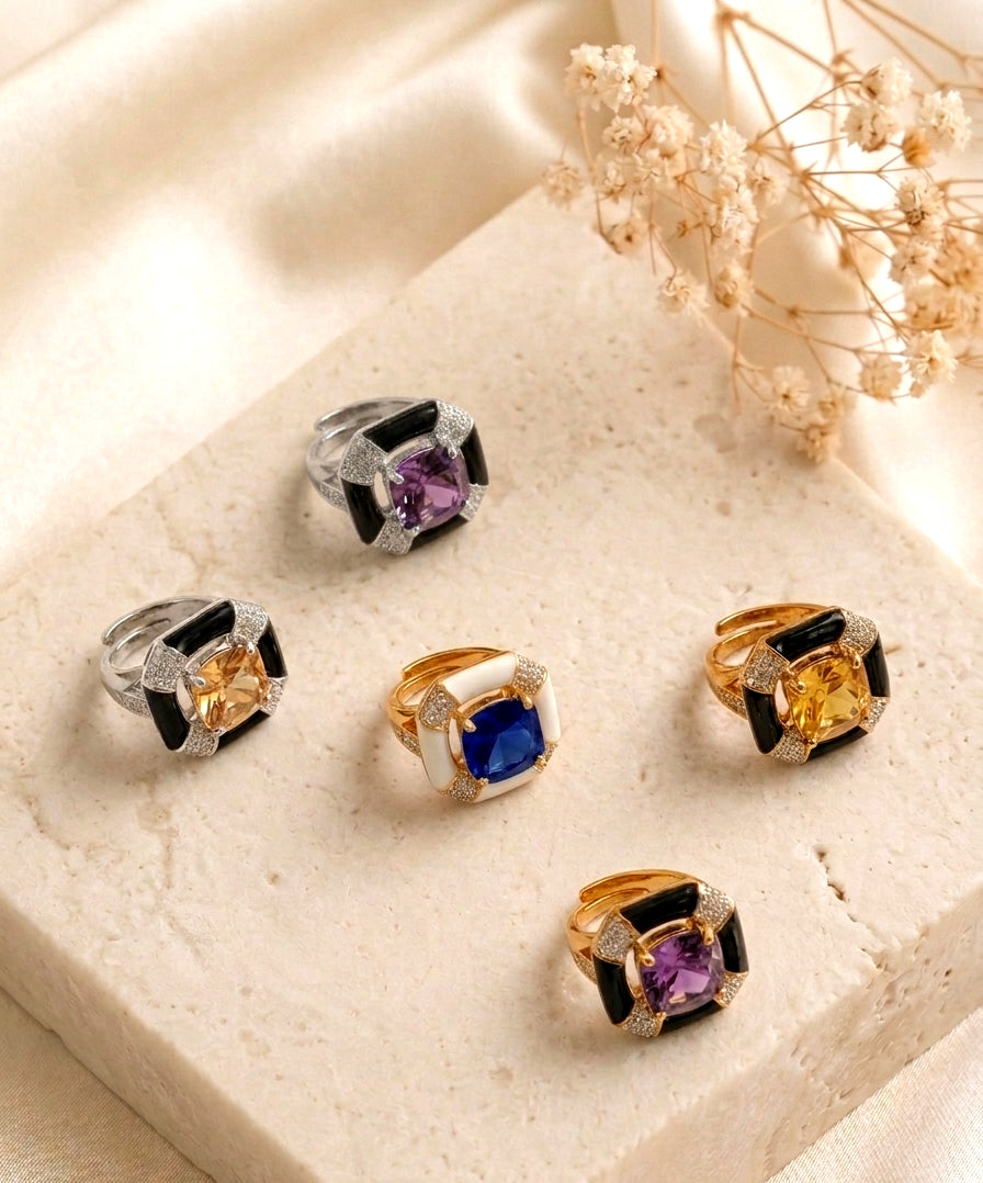 Five square-shaped rings with colorful gemstones and black enamel details are arranged on a beige stone slab with dried flowers in the background.