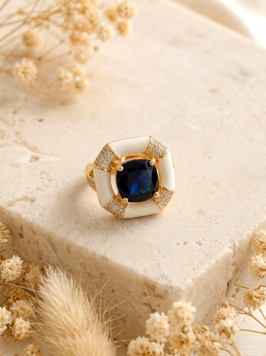 A gold ring with a large square blue gemstone and white enamel inlays, accented with small clear crystals, displayed on a beige stone block with dried flowers.