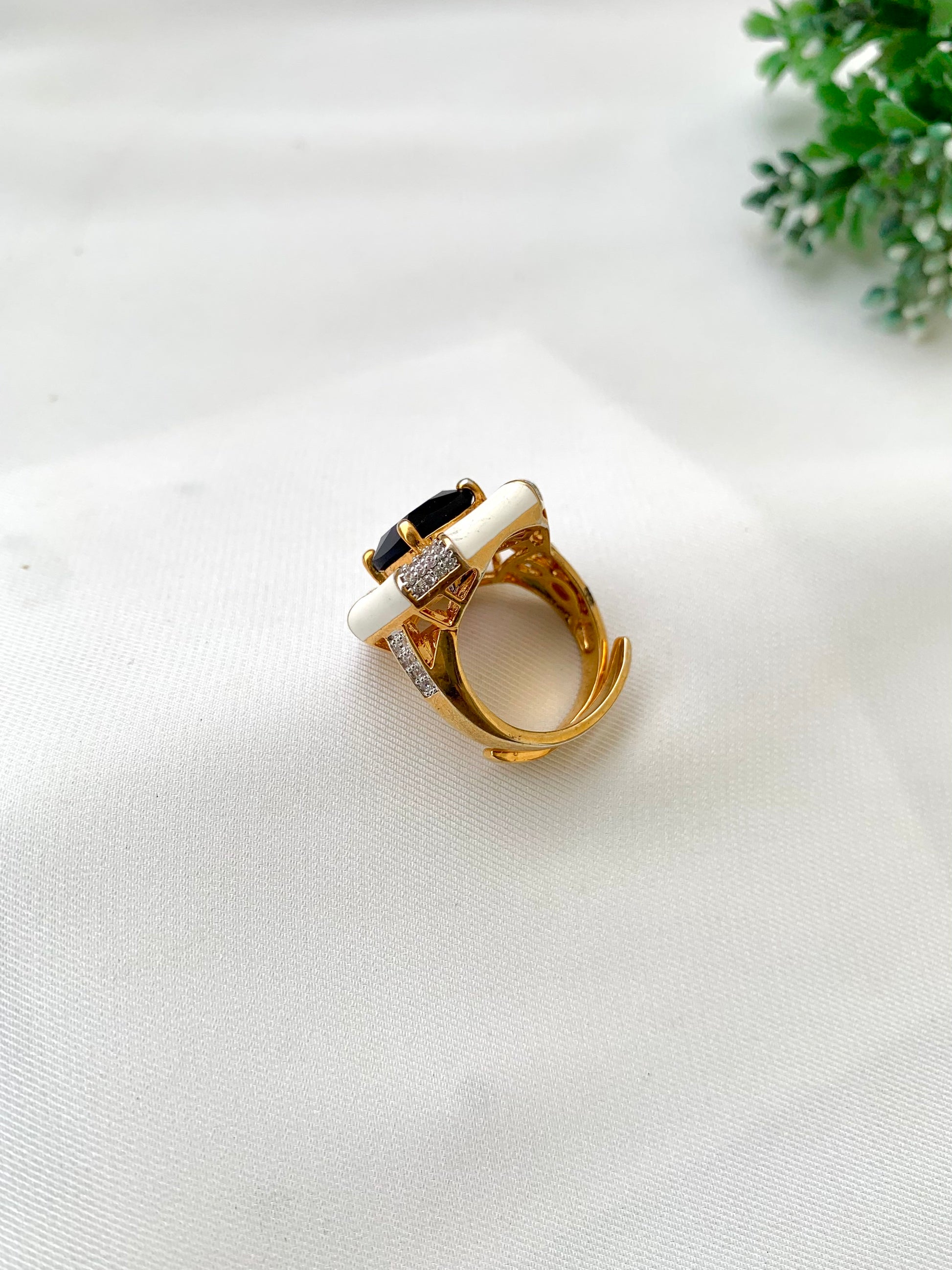 A gold ring with a white inlay and a black oval center stone, accented with small clear crystals, resting on a white textured fabric surface with a small green plant in the background.