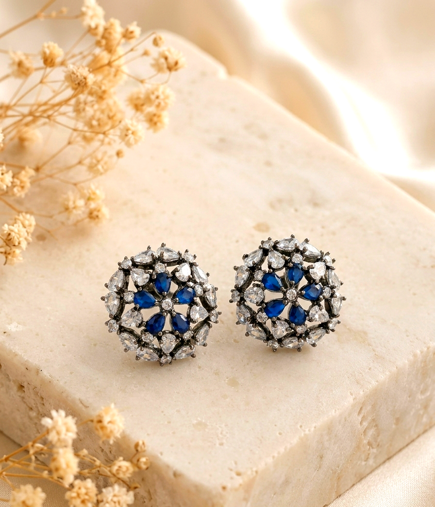 A pair of round stud earrings with a floral design, featuring deep blue gemstones in the center surrounded by clear crystals in a silver-toned setting, displayed on a beige stone block with dried flowers.
