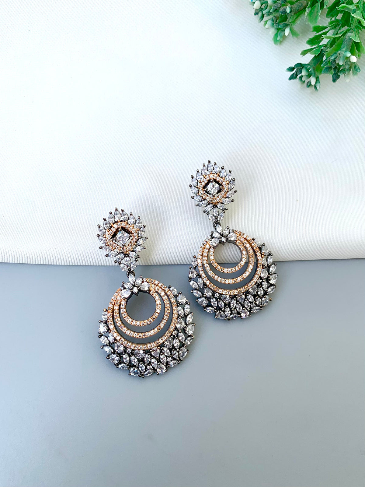 A pair of ornate chandelier earrings with silver-toned metal, rose gold-toned accents, and clear rhinestones, displayed on a split white and gray surface with a small green plant in the corner.