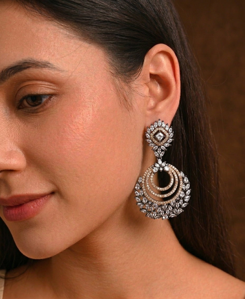 A close-up of a woman wearing a large, ornate silver-toned dangle earring with intricate crystal and gold-toned detailing, set against a warm brown background.