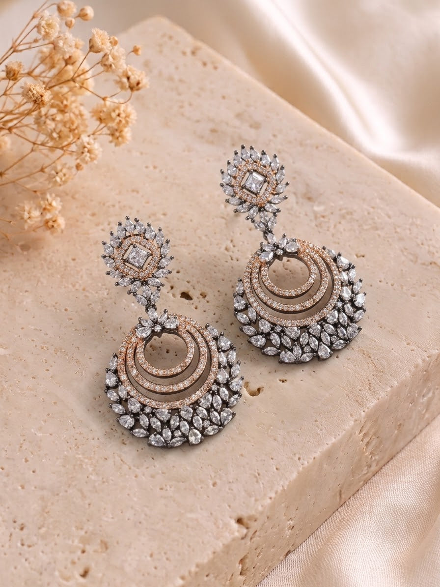 A pair of ornate chandelier earrings with silver-toned metal, rose gold-toned accents, and clear rhinestones, displayed on a split white and gray surface with a small green plant in the corner.