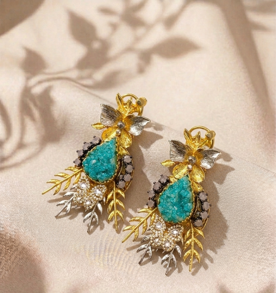 A pair of ornate gold-toned drop earrings with turquoise blue teardrop stones, silver-toned leaf details, and small clear stones, displayed on a beige fabric background with soft leaf shadows.
