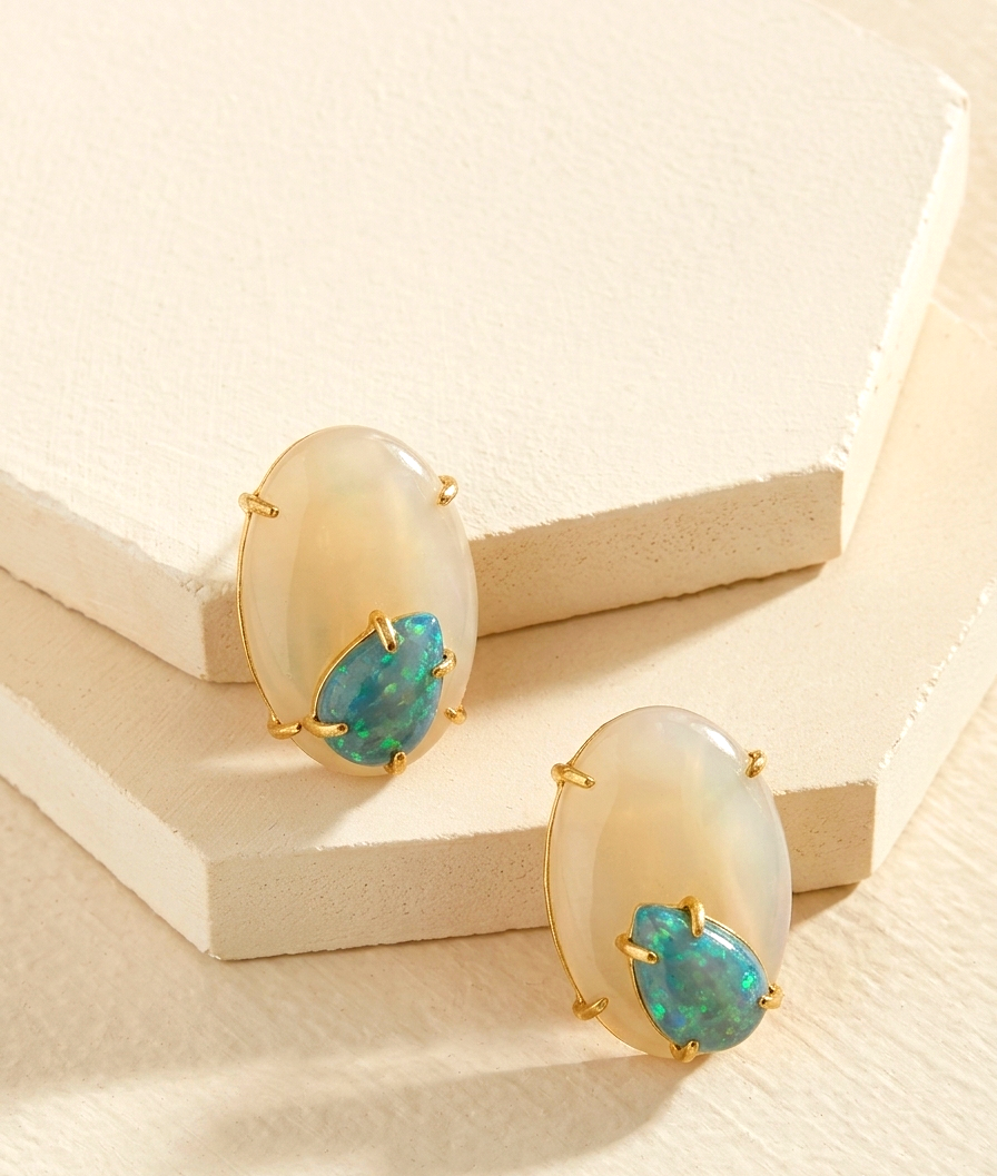A pair of gold-tone stud earrings with oval milky white cabochon stones and a smaller marquise-cut blue-green opal inset, displayed on a textured cream geometric block.