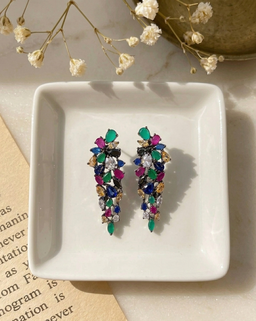 A pair of colorful statement dangle earrings with faceted gemstone-like pieces in green, pink, blue, purple, and gold, displayed on a small white square dish with dried flowers and a book in the background.