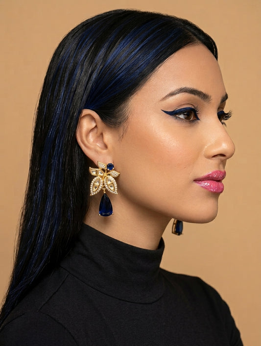 A woman in profile wearing a gold bow-shaped earring with dark blue teardrop gemstones and clear crystal accents, against a warm beige background.