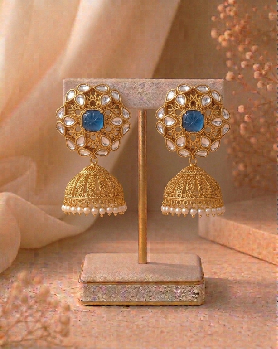A pair of ornate gold-toned jhumka earrings with blue square stones and white pearl-like accents, displayed on a beige jewelry stand against a warm beige background.