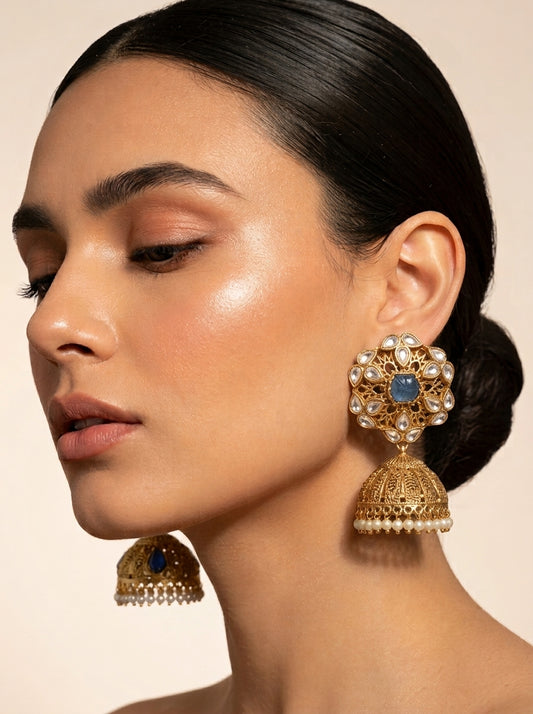 A close-up of a woman wearing ornate gold-toned Indian jhumka earrings with white faux pearls and blue stone accents, shown against a warm beige background.