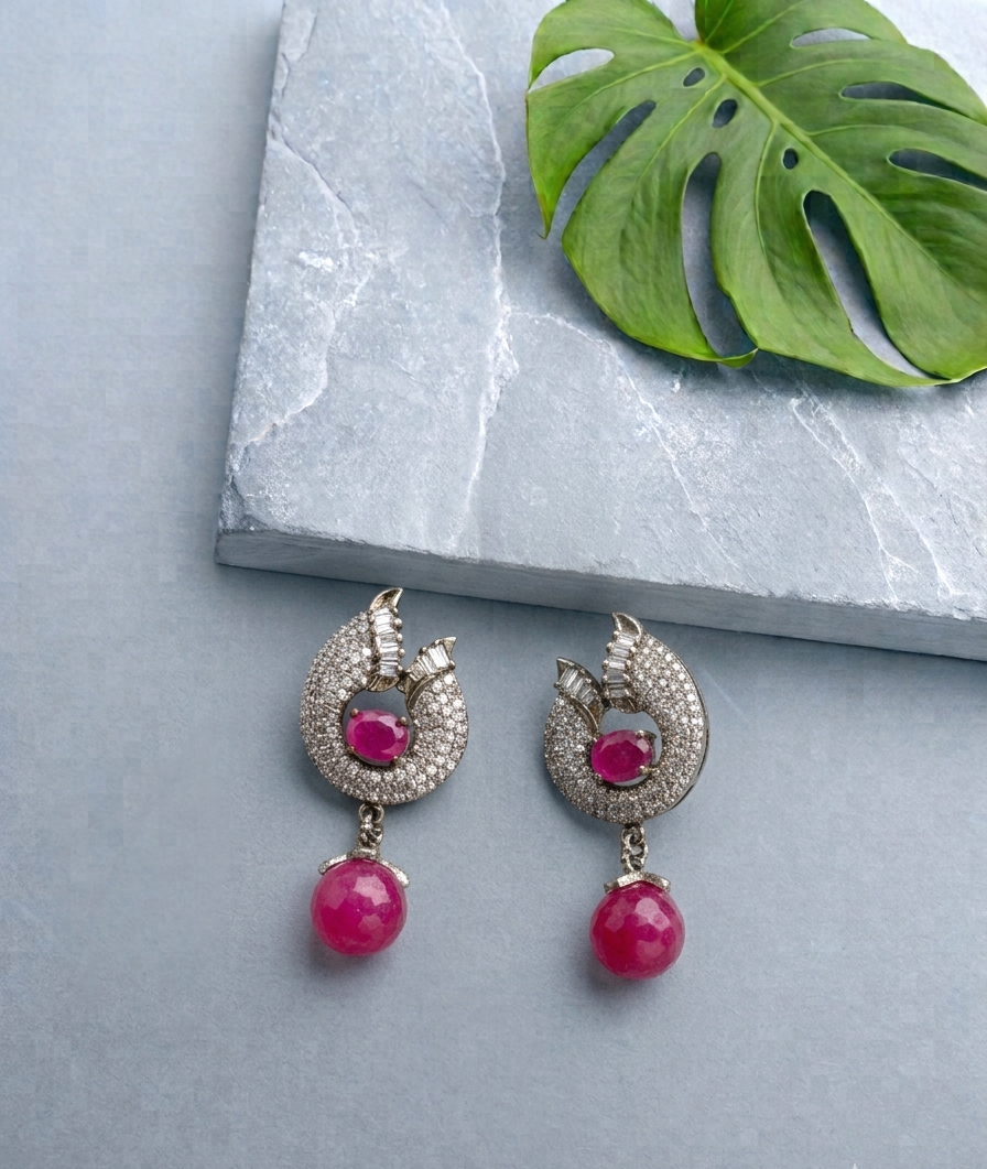 A pair of silver-toned drop earrings with a crescent-shaped pavé setting, a central round pink gemstone, and a dangling round pink bead, displayed on a light gray stone slab with a green monstera leaf.