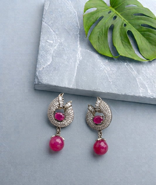 A pair of silver-toned drop earrings with a crescent-shaped pavé setting, a central round pink gemstone, and a dangling round pink bead, displayed on a light gray stone slab with a green monstera leaf.