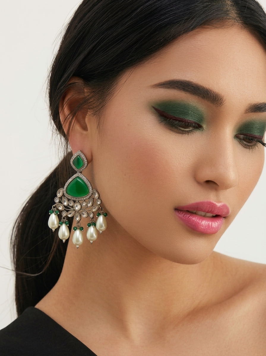 A close-up of a woman wearing a large green and silver teardrop-shaped chandelier earring with dangling pearl-like beads, against a clean light background.