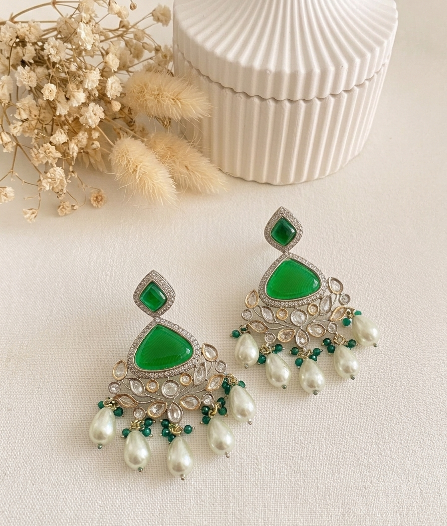 A pair of silver-toned chandelier earrings with vibrant green teardrop-shaped stones, small green accent beads, and dangling white pearl-like drops, styled on a light beige surface with dried flowers and a ribbed white container in the background.