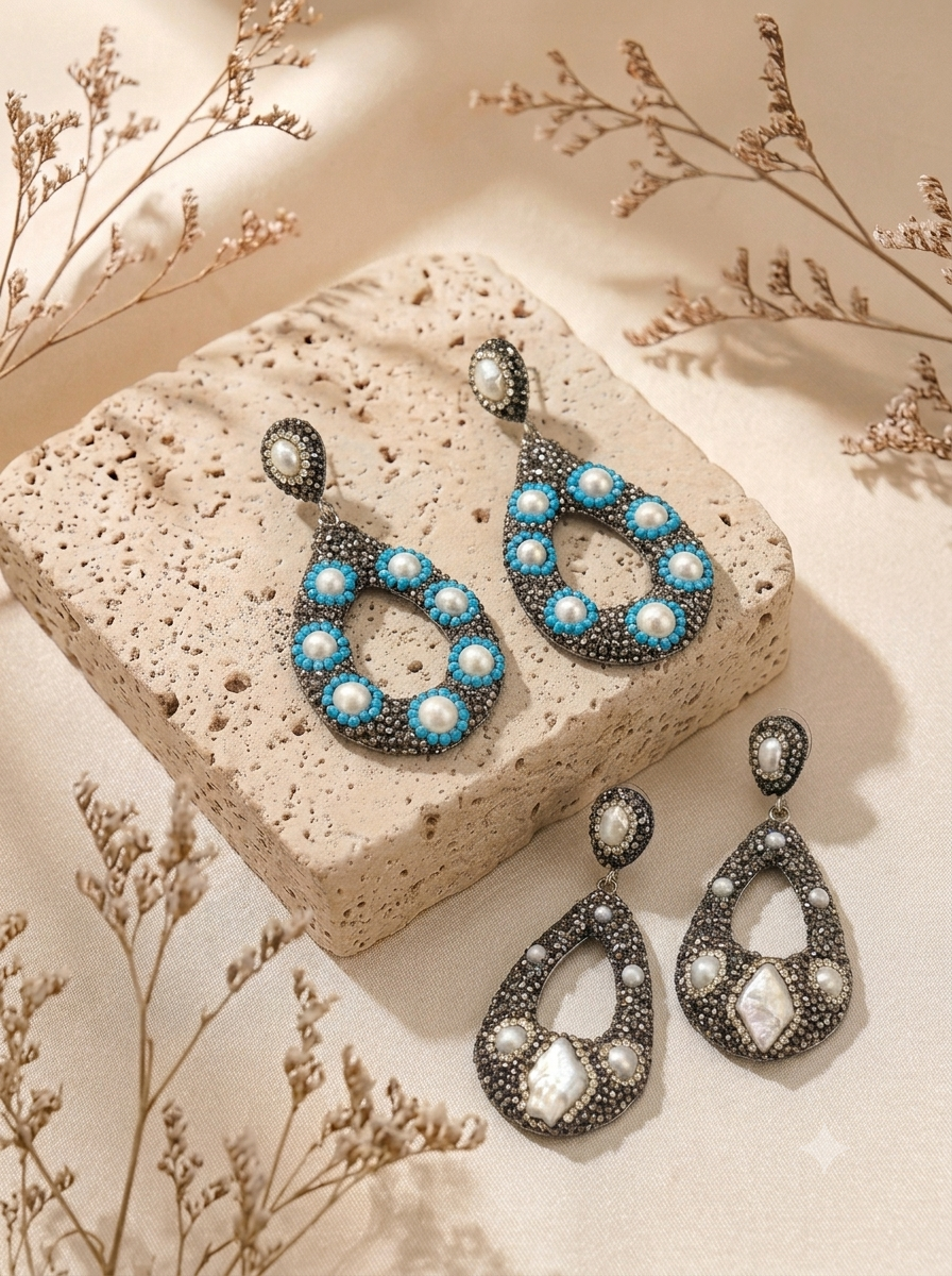 A pair of teardrop-shaped drop earrings displayed on a textured beige stone block with dried floral stems. The earrings feature silver-toned metal settings adorned with turquoise blue beads and white faux pearls, with larger clear rhinestones in the lower teardrop sections.