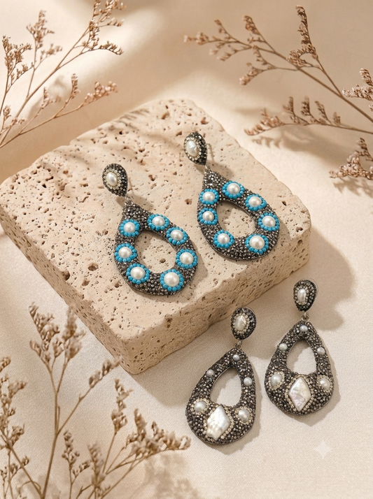 A pair of teardrop-shaped drop earrings displayed on a textured beige stone block with dried floral stems. The earrings feature silver-toned metal settings adorned with turquoise blue beads and white faux pearls, with larger clear rhinestones in the lower teardrop sections.