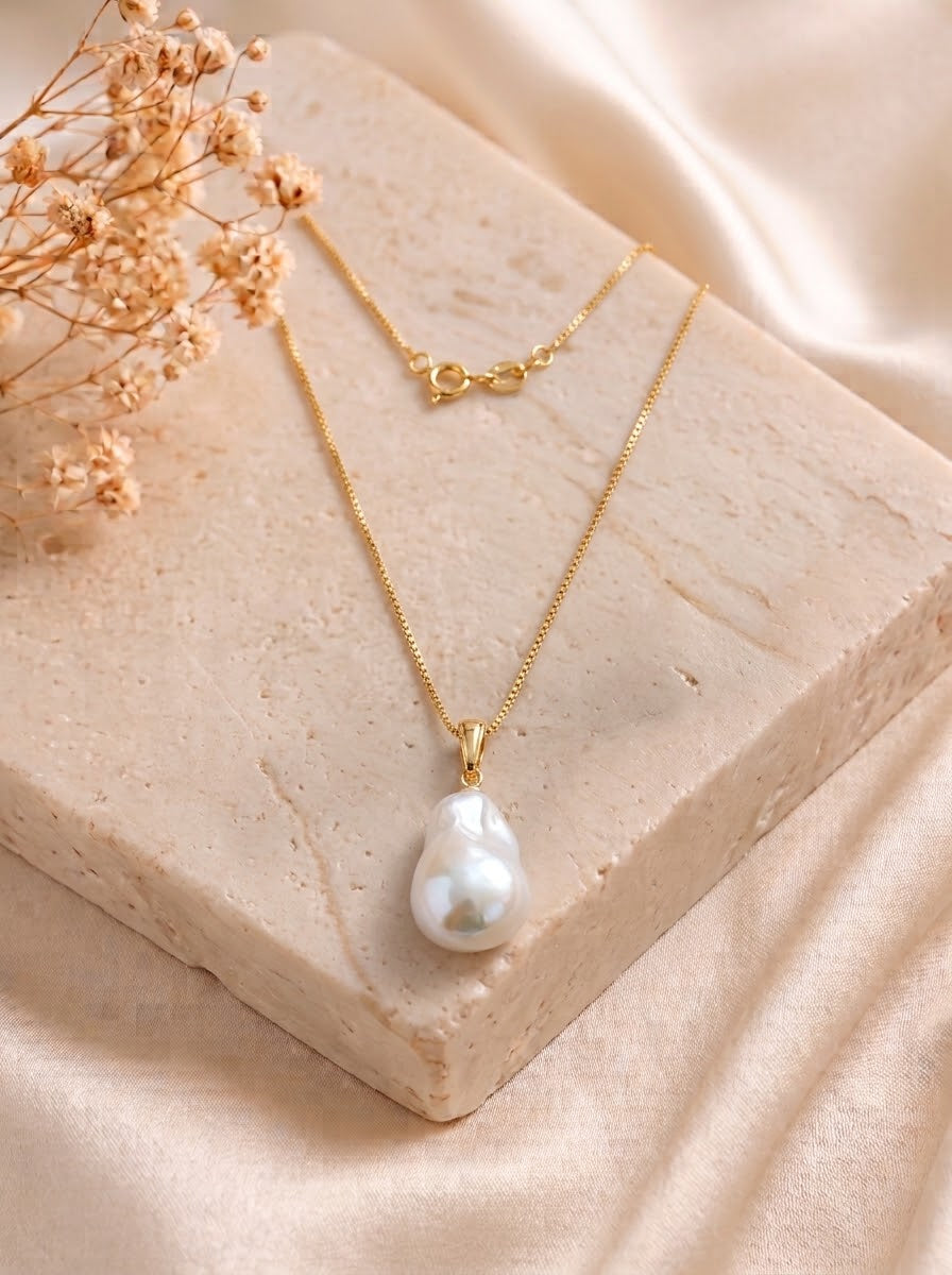 A gold chain necklace with a teardrop-shaped white baroque pearl pendant, displayed on a beige stone block with dried flowers and soft satin fabric.