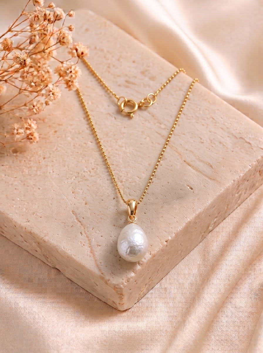 A gold chain necklace with a single irregular white pearl pendant, displayed on a beige stone block with dried flowers and soft satin fabric in the background.