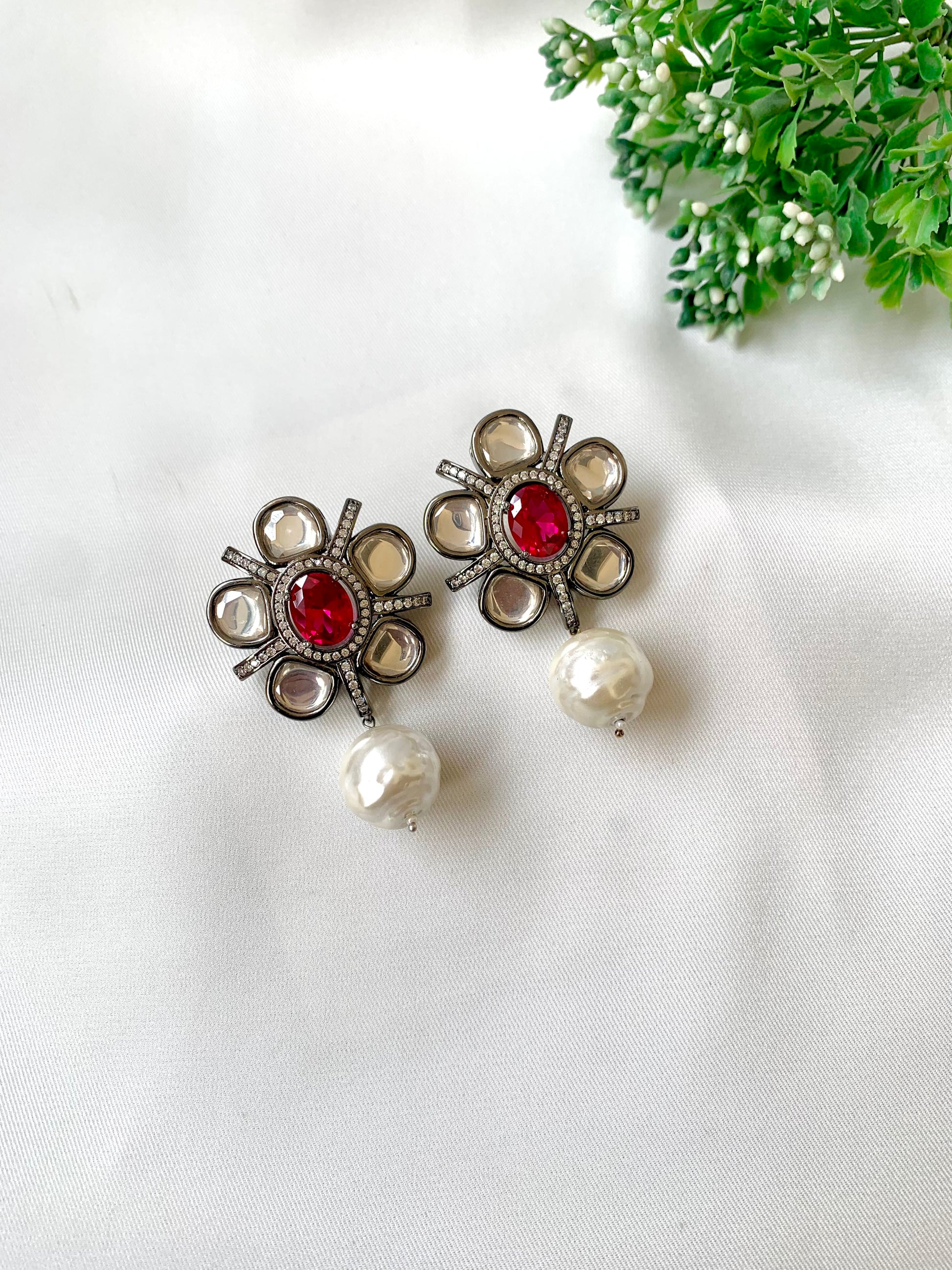 Pair of earrings with red gemstones and pearl-like beads on a white background with greenery.