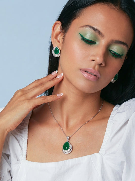 A close-up lifestyle shot of a model wearing a silver teardrop pendant necklace and matching drop earrings with green gemstones and clear accent stones, paired with a white top against a light blue background.