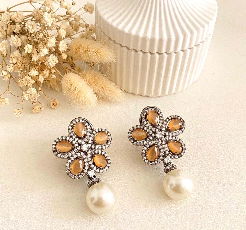 A pair of floral drop earrings with amber-colored teardrop stones, a halo of clear rhinestones, and a dangling white pearl, displayed on a light cream surface with dried flowers and a white ribbed box in the background.